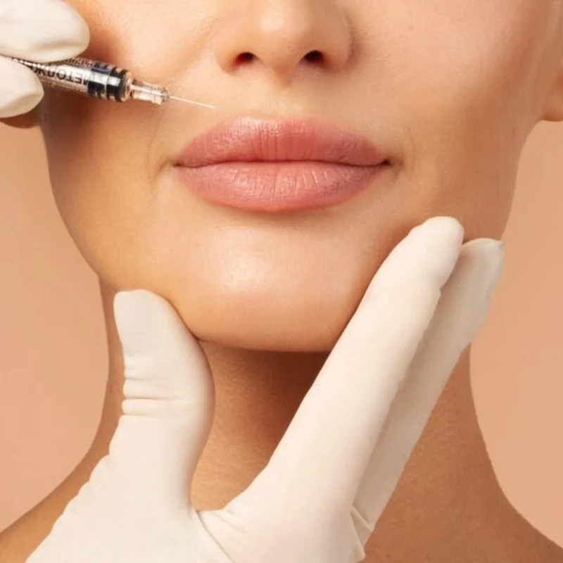 Gloved hands holding a patient’s chin while a syringe is positioned near the lips for dermal filler